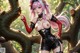 A woman with pink hair and a black outfit stands in a forest setting.